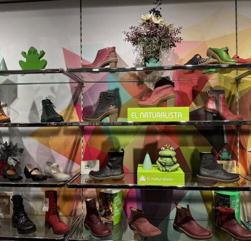 Regal mit bunten Schuhen und Pflanzen, bunte geometrische Wand, El Naturalista-Logo in Grün.