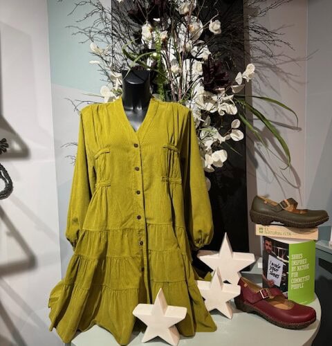 Schaufensterpuppe mit gelbem Kleid, neutraler Ausdruck, schwarze Perücke, umgeben von Schuhen.