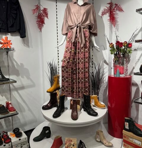 Eine Schaufensterpuppe trägt ein rosa Hemd und ein gemustertes Kleid, umgeben von bunten Schuhen.