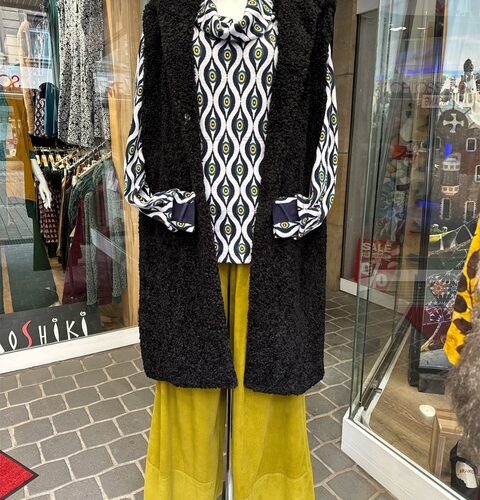Schaufenster-Mannequin in Retro-Outfit mit Augenprint-Hemd, schwarzer Weste und gelber Hose.