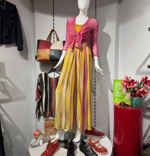 Eine Schaufensterpuppe im gelben Kleid und pinker Jacke, umgeben von Schuhen und Taschen.