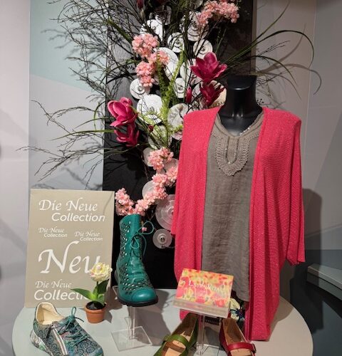 Schaufensterpuppe mit rosa Strickjacke, umgeben von Blumen und bunten Schuhen.