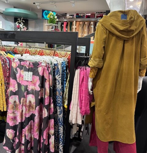 Schaufensterpuppe in einem senfgelben Mantel mit pinker Hose neben bunten, floralen Röcken.