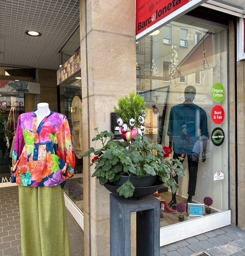 Schaufenster mit bunter Bluse und grüner Rock auf Mannequin, Pflanzen im Vordergrund.
