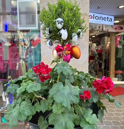 Pflanzgefäß mit bunten Eiern, grünen Blättern und roten Blüten in Einkaufsstraße.