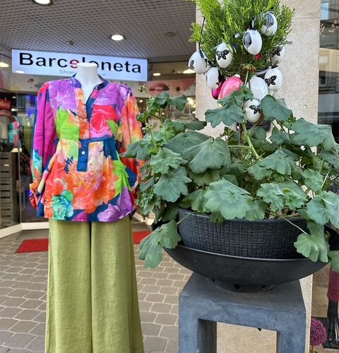 Schaufensterpuppe mit bunter Bluse und grüner Hose neben bepflanztem Blumentopf vor Geschäft.