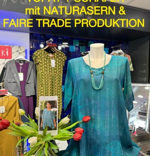 Schaufenster mit türkisfarbenem Kleid, Modeplakat, Blumensträußen und textilen Produkten im Hintergrund.